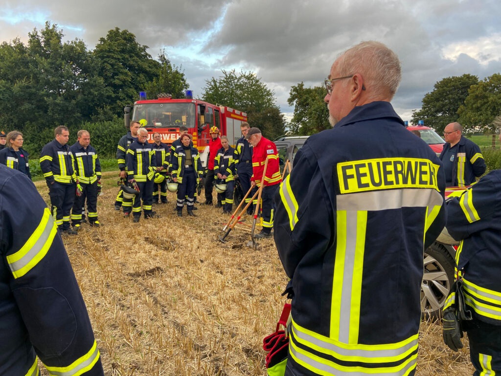 Vor einem Feuerwehrauto stehen zahlreiche Feuerwehrleute in Einsatzkleidung im Kreis. Ein Experte für Vegetationsbrandbekämpfung erklärt einige Einsatzmittel zur effektiven Brandbekämpfung.