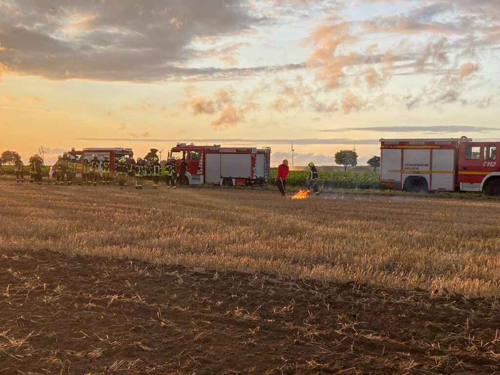 Das Bild zeigt drei Fahrzeuge am Feldrand. Einsatzkräfte der Feuerwehr stehen auf einem Stoppelfeld und beobachten einen Brand.